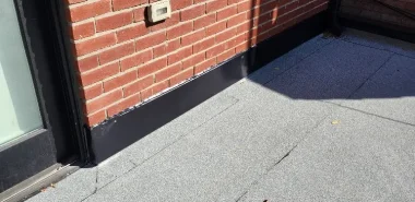 Grey flat roofing membrane with black metal flashing installed along a brick exterior wall and doorway.
