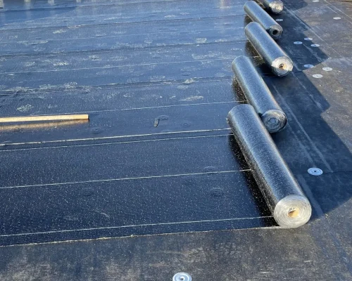 Close-up of a flat roof in the process of new construction roofing for commercial properties. Several rolls of waterproofing membrane are partially unrolled on the dark surface. Tools and wooden beams are visible in the background, along with a grassy area and trees. Dominion Roofing | BRAMPTON | BURLINGTON | ETOBICOKE | MARKHAM | MISSISSAUGA | NORTH YORK | OAKVILLE | RICHMOND HILL | Scarborough | TORONTO | VAUGHAN | dominionroofing.com | Phone Number: 416-789-0601