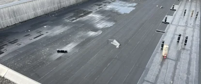 Workers are installing a black waterproof membrane on a large, flat commercial rooftop, showcasing top-tier commercial roof replacement solutions. One worker operates machinery as pipes protrude from the surface, surrounded by trees and industrial structures. Dominion Roofing | BRAMPTON | BURLINGTON | ETOBICOKE | MARKHAM | MISSISSAUGA | NORTH YORK | OAKVILLE | RICHMOND HILL | Scarborough | TORONTO | VAUGHAN | dominionroofing.com | Phone Number: 416-789-0601