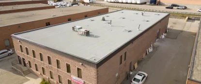 Aerial view of a large, rectangular, flat-roofed industrial building with brick walls. Several parked cars are visible nearby, and the surrounding area has additional industrial structures and greenery in the background. Dominion Roofing | BRAMPTON | BURLINGTON | ETOBICOKE | MARKHAM | MISSISSAUGA | NORTH YORK | OAKVILLE | RICHMOND HILL | Scarborough | TORONTO | VAUGHAN | dominionroofing.com | Phone Number: 416-789-0601