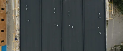 Aerial view of a flat, black roof with multiple vent pipes, surrounded by scaffolding and construction materials. The site seems to be part of new construction roofing for commercial properties, with wooden structures and trees visible around the area. Dominion Roofing | BRAMPTON | BURLINGTON | ETOBICOKE | MARKHAM | MISSISSAUGA | NORTH YORK | OAKVILLE | RICHMOND HILL | Scarborough | TORONTO | VAUGHAN | dominionroofing.com | Phone Number: 416-789-0601