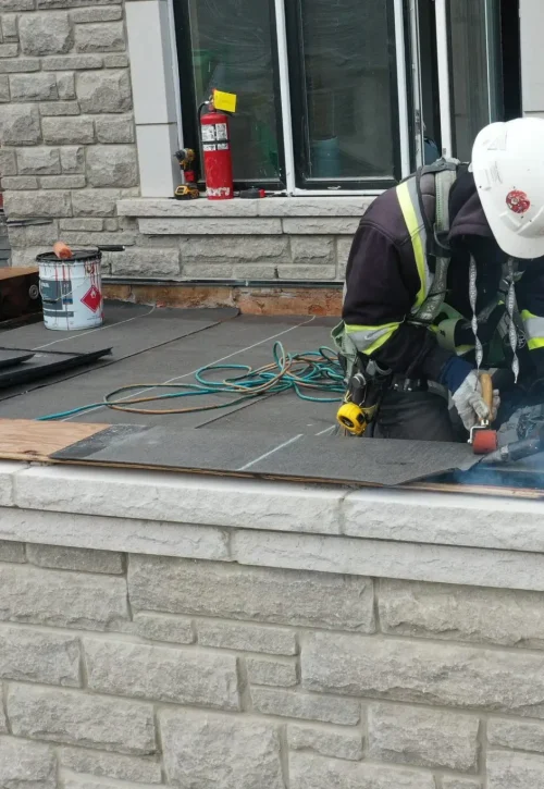 A construction worker in safety gear is using tools to work on the residential flat roofing of a building. The wall is made of brick and stone, and theres a window above. Safety ropes and equipment are visible around the worker. Dominion Roofing | BRAMPTON | BURLINGTON | ETOBICOKE | MARKHAM | MISSISSAUGA | NORTH YORK | OAKVILLE | RICHMOND HILL | Scarborough | TORONTO | VAUGHAN | dominionroofing.com | Phone Number: 416-789-0601