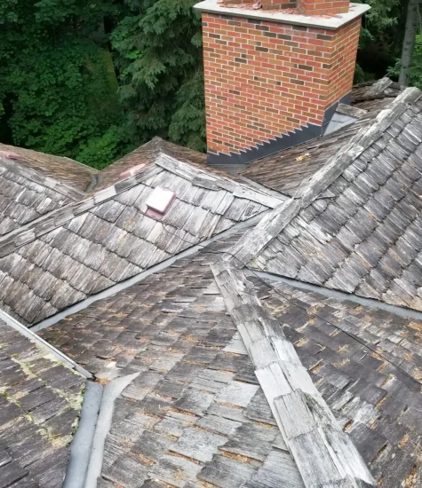 A worn wooden shingle roof, crafted with specialty residential roofing techniques, boasts multiple peaks and a red brick chimney. The aging shingles reveal weathered charm. Encircled by green leafy trees, it nestles beautifully in its natural backdrop. Dominion Roofing | BRAMPTON | BURLINGTON | ETOBICOKE | MARKHAM | MISSISSAUGA | NORTH YORK | OAKVILLE | RICHMOND HILL | Scarborough | TORONTO | VAUGHAN | dominionroofing.com | Phone Number: 416-789-0601