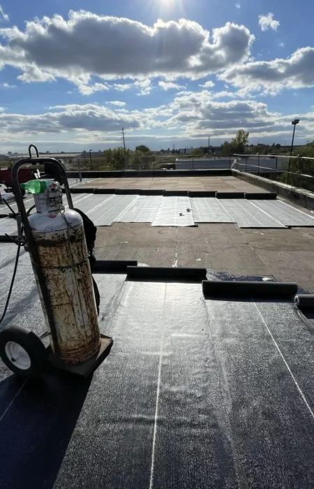 Construction workers on a flat rooftop are expertly installing black waterproofing membranes as part of comprehensive commercial roof replacement solutions. A gas tank with a hose lies nearby under the partly cloudy sky, with trees visible in the distance. Dominion Roofing | BRAMPTON | BURLINGTON | ETOBICOKE | MARKHAM | MISSISSAUGA | NORTH YORK | OAKVILLE | RICHMOND HILL | Scarborough | TORONTO | VAUGHAN | dominionroofing.com | Phone Number: 416-789-0601