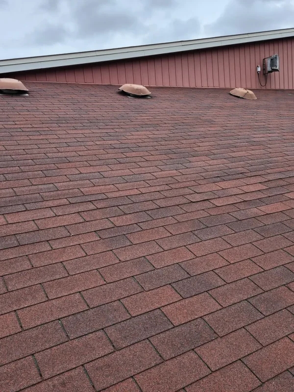The image depicts a residential shingle roofing scene, featuring a sloped roof with reddish-brown asphalt shingles. Four circular vents punctuate the roofline, and the building is finished with a beige trim against an overcast sky filled with gray clouds. Dominion Roofing | BRAMPTON | BURLINGTON | ETOBICOKE | MARKHAM | MISSISSAUGA | NORTH YORK | OAKVILLE | RICHMOND HILL | Scarborough | TORONTO | VAUGHAN | dominionroofing.com | Phone Number: 416-789-0601