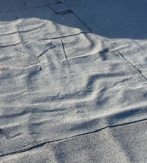 A rooftop with uneven, wrinkled gray asphalt shingles, typical of residential flat roofing, shows signs of wear and exposure to sunlight. Shadows accentuate the texture and irregularities in the surface. Dominion Roofing | BRAMPTON | BURLINGTON | ETOBICOKE | MARKHAM | MISSISSAUGA | NORTH YORK | OAKVILLE | RICHMOND HILL | Scarborough | TORONTO | VAUGHAN | dominionroofing.com | Phone Number: 416-789-0601