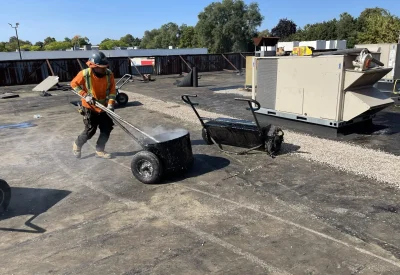 A construction worker in an orange vest and helmet expertly applies tar on a flat roof with a wheeled tar bucket, part of our advanced Commercial Roof Replacement Solutions. The roof is scattered with gravel and equipment, while trees stand tall under the clear sky. Dominion Roofing | BRAMPTON | BURLINGTON | ETOBICOKE | MARKHAM | MISSISSAUGA | NORTH YORK | OAKVILLE | RICHMOND HILL | Scarborough | TORONTO | VAUGHAN | dominionroofing.com | Phone Number: 416-789-0601