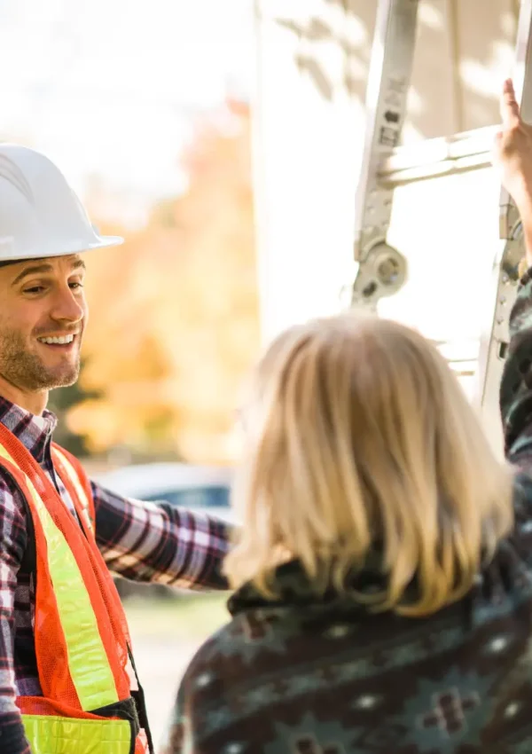 A construction worker in a safety vest and helmet smiles while talking to a woman. She points at a ladder leaning against a house. Autumn leaves are visible in the background. Dominion Roofing | BRAMPTON | BURLINGTON | ETOBICOKE | MARKHAM | MISSISSAUGA | NORTH YORK | OAKVILLE | RICHMOND HILL | Scarborough | TORONTO | VAUGHAN | dominionroofing.com | Phone Number: 416-789-0601