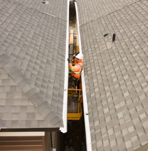 A worker in an orange safety vest and white helmet stands on a mobile platform, elevated near the roofline of a new construction home. Positioned between two gray-roofed buildings, they expertly handle roofing materials or guttering equipment, ensuring every detail aligns perfectly. Dominion Roofing | BRAMPTON | BURLINGTON | ETOBICOKE | MARKHAM | MISSISSAUGA | NORTH YORK | OAKVILLE | RICHMOND HILL | Scarborough | TORONTO | VAUGHAN | dominionroofing.com | Phone Number: 416-789-0601