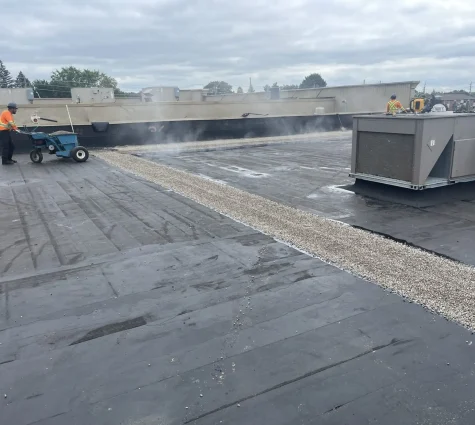 Workers on a flat rooftop are using equipment to apply a layer of gravel over the black surface, part of the new construction roofing for commercial properties. Steam or dust rises from the machinery, with buildings and trees visible in the background against a cloudy sky. Dominion Roofing | BRAMPTON | BURLINGTON | ETOBICOKE | MARKHAM | MISSISSAUGA | NORTH YORK | OAKVILLE | RICHMOND HILL | Scarborough | TORONTO | VAUGHAN | dominionroofing.com | Phone Number: 416-789-0601