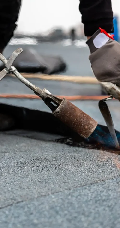 A person expertly uses a propane torch to apply and seal a roll of roofing material onto a residential flat roof. The flame flickers visibly as they work, wearing gloves and sturdy work boots for safety. Dominion Roofing | BRAMPTON | BURLINGTON | ETOBICOKE | MARKHAM | MISSISSAUGA | NORTH YORK | OAKVILLE | RICHMOND HILL | Scarborough | TORONTO | VAUGHAN | dominionroofing.com | Phone Number: 416-789-0601