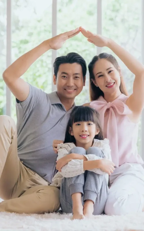 A family sitting on a soft carpet in front of large windows with trees outside. The parents form a roof shape with their arms above their smiling daughter, who is seated between them, hugging her knees. Dominion Roofing | BRAMPTON | BURLINGTON | ETOBICOKE | MARKHAM | MISSISSAUGA | NORTH YORK | OAKVILLE | RICHMOND HILL | Scarborough | TORONTO | VAUGHAN | dominionroofing.com | Phone Number: 416-789-0601