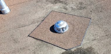 A flat rooftop with a metal drain cover in the center, surrounded by a patch of differently colored roofing material and a dark stain nearby. A vent pipe and a parapet wall are visible in the background. Dominion Roofing | BRAMPTON | BURLINGTON | ETOBICOKE | MARKHAM | MISSISSAUGA | NORTH YORK | OAKVILLE | RICHMOND HILL | Scarborough | TORONTO | VAUGHAN | dominionroofing.com | Phone Number: 416-789-0601