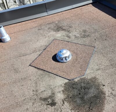 A flat rooftop with a metal drain cover in the center, surrounded by a patch of differently colored roofing material and a dark stain nearby. A vent pipe and a parapet wall are visible in the background. Dominion Roofing | BRAMPTON | BURLINGTON | ETOBICOKE | MARKHAM | MISSISSAUGA | NORTH YORK | OAKVILLE | RICHMOND HILL | Scarborough | TORONTO | VAUGHAN | dominionroofing.com | Phone Number: 416-789-0601
