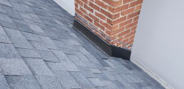 Gray asphalt shingles on a sloped roof with a red brick chimney and metal flashing at the base, next to a white wall. Dominion Roofing | BRAMPTON | BURLINGTON | ETOBICOKE | MARKHAM | MISSISSAUGA | NORTH YORK | OAKVILLE | RICHMOND HILL | Scarborough | TORONTO | VAUGHAN | dominionroofing.com | Phone Number: 416-789-0601