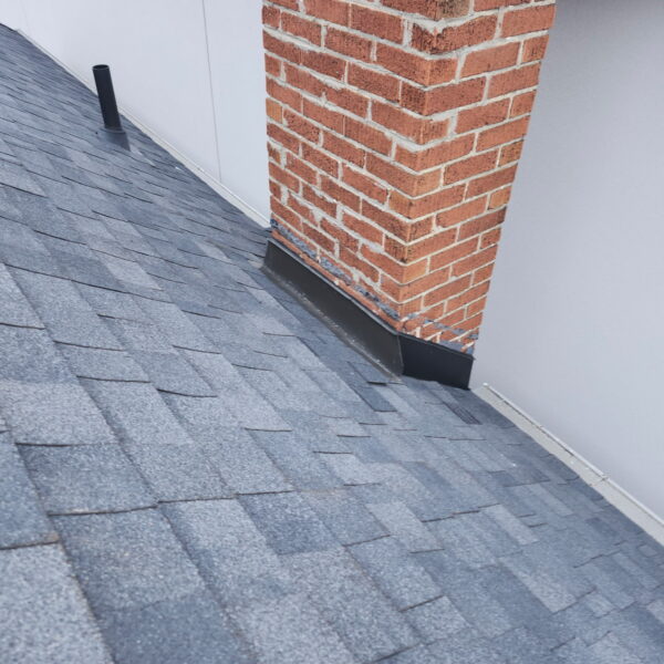 Gray asphalt shingles on a sloped roof with a red brick chimney and metal flashing at the base, next to a white wall. Dominion Roofing | BRAMPTON | BURLINGTON | ETOBICOKE | MARKHAM | MISSISSAUGA | NORTH YORK | OAKVILLE | RICHMOND HILL | Scarborough | TORONTO | VAUGHAN | dominionroofing.com | Phone Number: 416-789-0601
