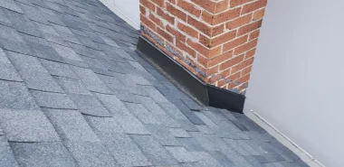 Gray asphalt shingles on a sloped roof with a red brick chimney and metal flashing at the base, next to a white wall. Dominion Roofing | BRAMPTON | BURLINGTON | ETOBICOKE | MARKHAM | MISSISSAUGA | NORTH YORK | OAKVILLE | RICHMOND HILL | Scarborough | TORONTO | VAUGHAN | dominionroofing.com | Phone Number: 416-789-0601