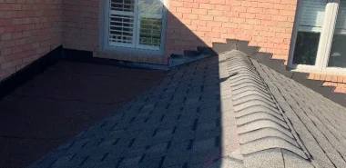Residential roofing system featuring grey asphalt shingles, a skylight, roof ridges and a flat roof section adjoining brick walls.