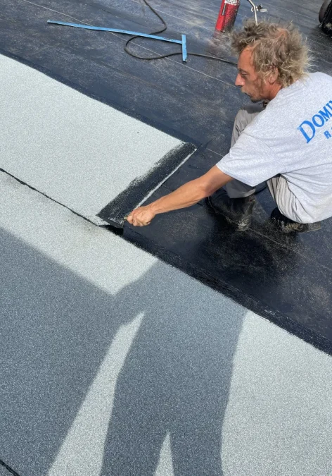 A person kneels on a commercial flat roof, applying adhesive between gray roofing sheets. Dressed in a gray shirt, they have tools nearby. The shadow of another person taking the photo is visible on the roofing material. Dominion Roofing | BRAMPTON | BURLINGTON | ETOBICOKE | MARKHAM | MISSISSAUGA | NORTH YORK | OAKVILLE | RICHMOND HILL | Scarborough | TORONTO | VAUGHAN | dominionroofing.com | Phone Number: 416-789-0601