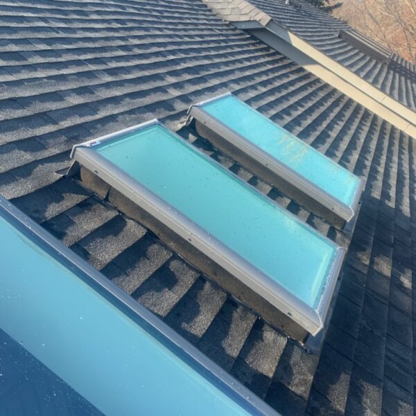 Three rectangular skylights set into a dark gray shingled roof, with trees and clear sky visible in the background. The skylights have a blue tint and are aligned in a row. Dominion Roofing | BRAMPTON | BURLINGTON | ETOBICOKE | MARKHAM | MISSISSAUGA | NORTH YORK | OAKVILLE | RICHMOND HILL | Scarborough | TORONTO | VAUGHAN | dominionroofing.com | Phone Number: 416-789-0601