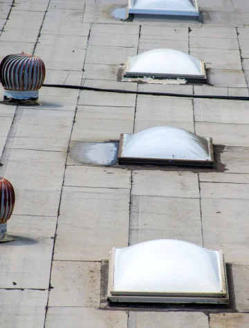 A flat rooftop designed for commercial roof inspection boasts several white, dome-shaped skylights and two striped, spherical ventilation turbines. A few cables discreetly traverse the grey surface, maintaining functionality and accessibility. Dominion Roofing | BRAMPTON | BURLINGTON | ETOBICOKE | MARKHAM | MISSISSAUGA | NORTH YORK | OAKVILLE | RICHMOND HILL | Scarborough | TORONTO | VAUGHAN | dominionroofing.com | Phone Number: 416-789-0601