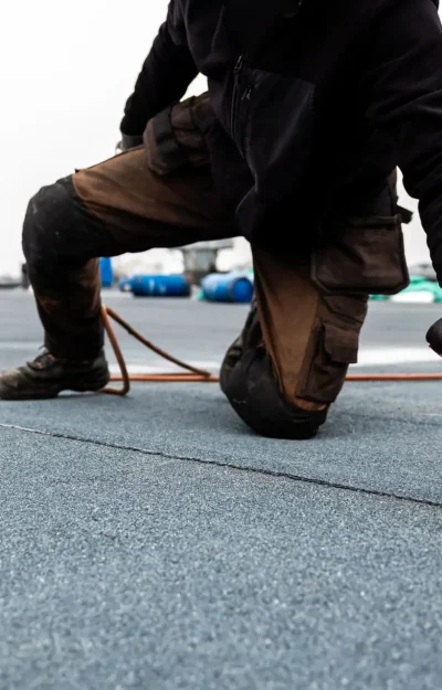 A person wearing work gloves is kneeling on a flat rooftop, engaged in a commercial roof inspection. With a tank and hose nearby, they diligently use their tool on the surface as the overcast sky looms above, trees standing quietly in the distance. Dominion Roofing | BRAMPTON | BURLINGTON | ETOBICOKE | MARKHAM | MISSISSAUGA | NORTH YORK | OAKVILLE | RICHMOND HILL | Scarborough | TORONTO | VAUGHAN | dominionroofing.com | Phone Number: 416-789-0601