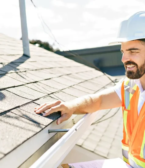 A construction worker, wearing a hard hat and an orange safety vest, conducts a residential roof inspection. Standing near the edge with a clipboard in hand, he carefully examines the shingles. Its a bright day, with trees and a building adding to the scenes backdrop. Dominion Roofing | BRAMPTON | BURLINGTON | ETOBICOKE | MARKHAM | MISSISSAUGA | NORTH YORK | OAKVILLE | RICHMOND HILL | Scarborough | TORONTO | VAUGHAN | dominionroofing.com | Phone Number: 416-789-0601