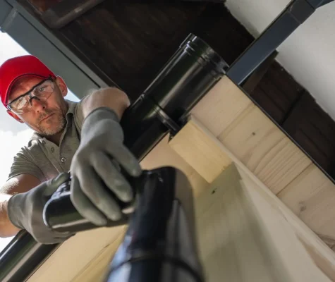 A person wearing a red cap, glasses, and gloves is installing or repairing a black gutter on a house. The photo is taken from below, looking up at the individual focused on their work. The sky is partly cloudy. Dominion Roofing | BRAMPTON | BURLINGTON | ETOBICOKE | MARKHAM | MISSISSAUGA | NORTH YORK | OAKVILLE | RICHMOND HILL | Scarborough | TORONTO | VAUGHAN | dominionroofing.com | Phone Number: 416-789-0601