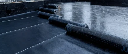 A construction site with several rolls of black roofing material laid out on a partially covered flat surface. The area shows black waterproof layers applied, and the open walls reveal the buildings structure. Dominion Roofing | BRAMPTON | BURLINGTON | ETOBICOKE | MARKHAM | MISSISSAUGA | NORTH YORK | OAKVILLE | RICHMOND HILL | Scarborough | TORONTO | VAUGHAN | dominionroofing.com | Phone Number: 416-789-0601