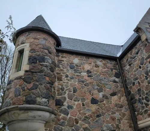 A stone building boasts a corner turret with a conical roof, showcasing the elegance of specialty commercial roofing solutions. Made of multicolored stones, its charm is enhanced by an overcast sky and trees peeking in from the left. A small window sits quaintly in the turret. Dominion Roofing | BRAMPTON | BURLINGTON | ETOBICOKE | MARKHAM | MISSISSAUGA | NORTH YORK | OAKVILLE | RICHMOND HILL | Scarborough | TORONTO | VAUGHAN | dominionroofing.com | Phone Number: 416-789-0601