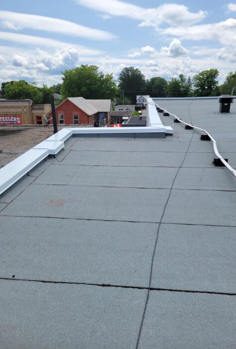 Flat rooftop with light gray covering, indicating recent roof repair, features cables and a metal edge. The background shows small red brick buildings and green trees under a partly cloudy sky. Dominion Roofing | BRAMPTON | BURLINGTON | ETOBICOKE | MARKHAM | MISSISSAUGA | NORTH YORK | OAKVILLE | RICHMOND HILL | Scarborough | TORONTO | VAUGHAN | dominionroofing.com | Phone Number: 416-789-0601