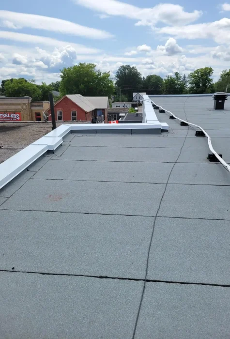 Flat rooftop with light gray covering, indicating recent roof repair, features cables and a metal edge. The background shows small red brick buildings and green trees under a partly cloudy sky. Dominion Roofing | BRAMPTON | BURLINGTON | ETOBICOKE | MARKHAM | MISSISSAUGA | NORTH YORK | OAKVILLE | RICHMOND HILL | Scarborough | TORONTO | VAUGHAN | dominionroofing.com | Phone Number: 416-789-0601