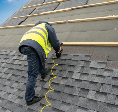 A worker in a yellow safety vest is installing asphalt shingles on a sloped roof. The worker uses a nail gun connected to a yellow air hose. The sky is partly cloudy. Dominion Roofing | BRAMPTON | BURLINGTON | ETOBICOKE | MARKHAM | MISSISSAUGA | NORTH YORK | OAKVILLE | RICHMOND HILL | Scarborough | TORONTO | VAUGHAN | dominionroofing.com | Phone Number: 416-789-0601