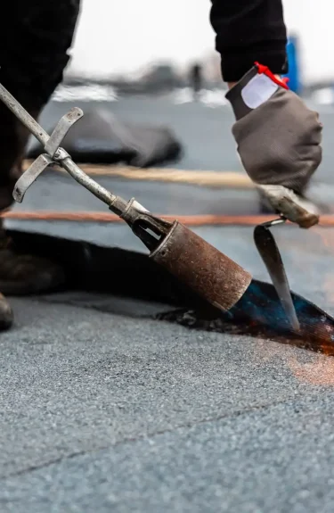 A construction worker, vital to the new construction roofing for commercial properties, dons gloves and boots. With precision, he uses a propane torch to heat and seal roofing material on a flat roof, the flame dancing as it ensures the surface adheres perfectly. Dominion Roofing | BRAMPTON | BURLINGTON | ETOBICOKE | MARKHAM | MISSISSAUGA | NORTH YORK | OAKVILLE | RICHMOND HILL | Scarborough | TORONTO | VAUGHAN | dominionroofing.com | Phone Number: 416-789-0601