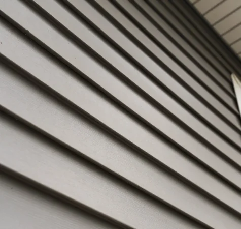 Close-up of a house exterior showcasing new construction with horizontal beige vinyl siding and a section of a white-framed window. The siding lines create a repetitive pattern, adding texture to the image. The eaves and part of the roof are visible at the top, highlighting modern roofing for homes. Dominion Roofing | BRAMPTON | BURLINGTON | ETOBICOKE | MARKHAM | MISSISSAUGA | NORTH YORK | OAKVILLE | RICHMOND HILL | Scarborough | TORONTO | VAUGHAN | dominionroofing.com | Phone Number: 416-789-0601