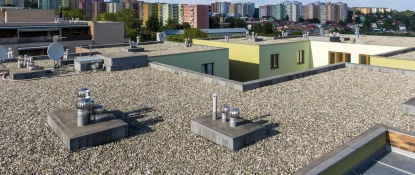 A flat rooftop covered with gravel, featuring several ventilation chimneys and circular vents. Surrounding buildings and a tree-filled landscape are visible in the background under a clear sky. Dominion Roofing | BRAMPTON | BURLINGTON | ETOBICOKE | MARKHAM | MISSISSAUGA | NORTH YORK | OAKVILLE | RICHMOND HILL | Scarborough | TORONTO | VAUGHAN | dominionroofing.com | Phone Number: 416-789-0601