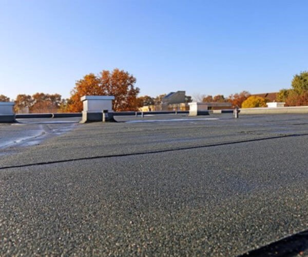 The flat rooftop, primed for roof repair, boasts a smooth asphalt surface under a clear blue sky. White ventilation units are scattered across the expanse, while autumn trees with orange leaves and a few buildings form the picturesque background. Dominion Roofing | BRAMPTON | BURLINGTON | ETOBICOKE | MARKHAM | MISSISSAUGA | NORTH YORK | OAKVILLE | RICHMOND HILL | Scarborough | TORONTO | VAUGHAN | dominionroofing.com | Phone Number: 416-789-0601
