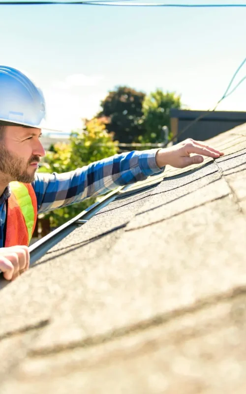 A construction worker wearing a hard hat and reflective vest inspects roof shingles on a sunny day. Engaged in commercial shingle roofing services, he ascends the ladder to assess the roofs surface condition, with trees and power lines providing a backdrop. Dominion Roofing | BRAMPTON | BURLINGTON | ETOBICOKE | MARKHAM | MISSISSAUGA | NORTH YORK | OAKVILLE | RICHMOND HILL | Scarborough | TORONTO | VAUGHAN | dominionroofing.com | Phone Number: 416-789-0601