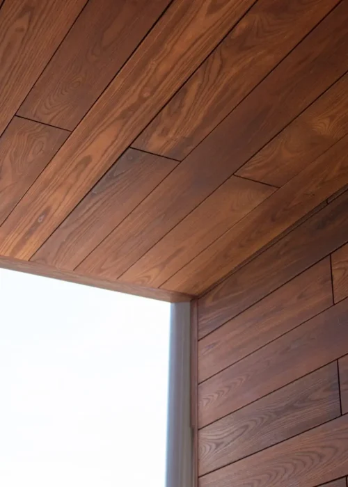 View of a modern architectural ceiling corner with wooden panels, seamlessly transitioning from the ceiling to the wall. The design, reminiscent of expert residential siding installation, features a warm finish and visible grain patterns for a minimalistic and clean aesthetic. Dominion Roofing | BRAMPTON | BURLINGTON | ETOBICOKE | MARKHAM | MISSISSAUGA | NORTH YORK | OAKVILLE | RICHMOND HILL | Scarborough | TORONTO | VAUGHAN | dominionroofing.com | Phone Number: 416-789-0601