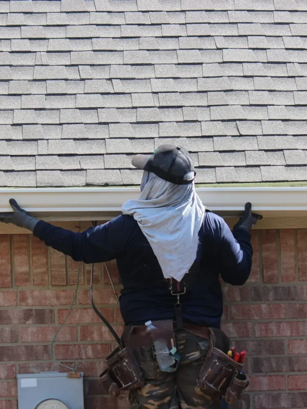A worker in a cap adjusts a gutter on a brick house, embodying professional Residential Eavestrough & Downpipe Services. They wear a long-sleeve shirt, gloves, and have fabric draped over their neck for sun protection. A tool belt with various tools is strapped around their waist. Dominion Roofing | BRAMPTON | BURLINGTON | ETOBICOKE | MARKHAM | MISSISSAUGA | NORTH YORK | OAKVILLE | RICHMOND HILL | Scarborough | TORONTO | VAUGHAN | dominionroofing.com | Phone Number: 416-789-0601