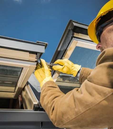 A construction worker wearing a yellow hard hat and gloves installs a skylight window on a roof. The worker uses a screwdriver to secure the frame under a clear blue sky. Dominion Roofing | BRAMPTON | BURLINGTON | ETOBICOKE | MARKHAM | MISSISSAUGA | NORTH YORK | OAKVILLE | RICHMOND HILL | Scarborough | TORONTO | VAUGHAN | dominionroofing.com | Phone Number: 416-789-0601